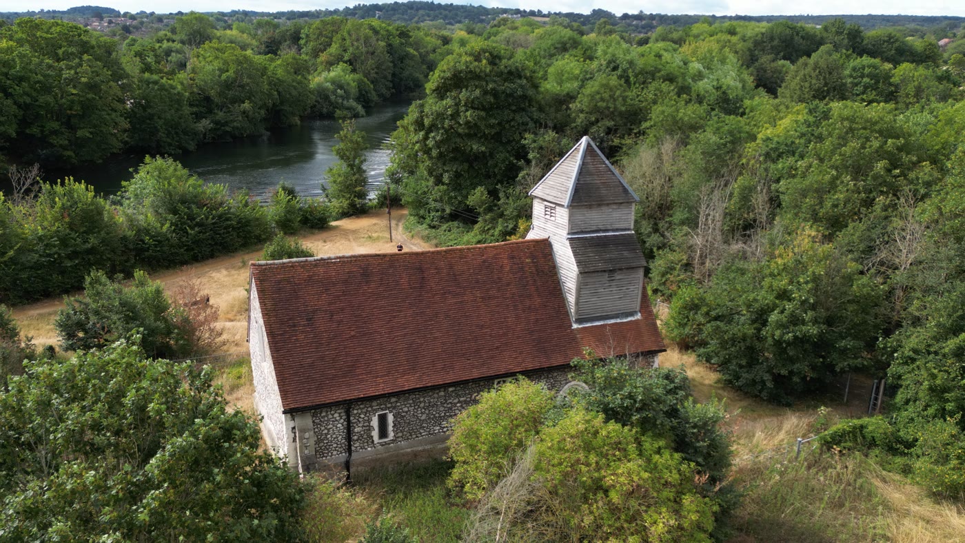 A flint church with a wooden tower seen from the air, the River Thames and wooded countryside behind it.
