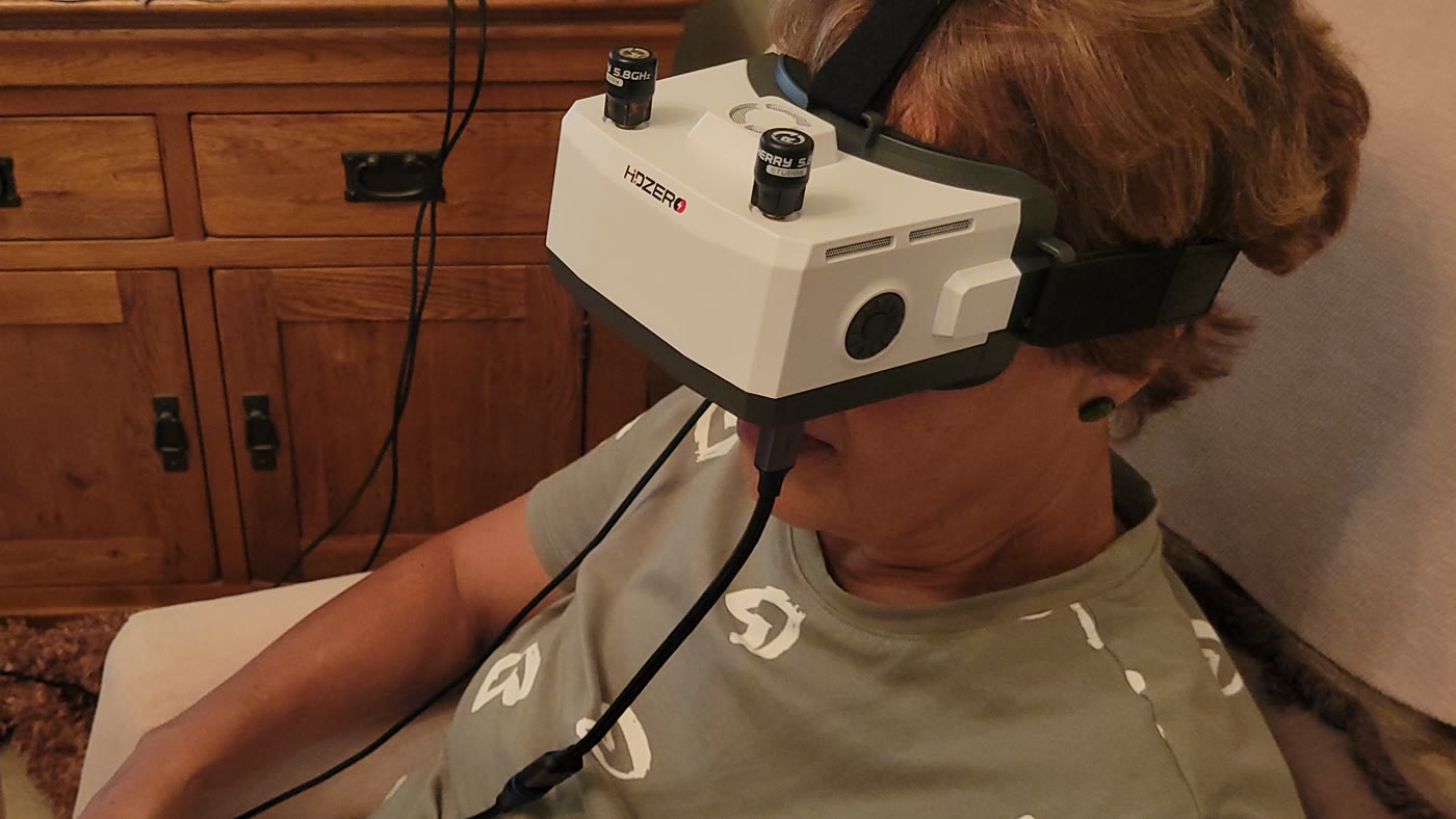 Box-style FPV goggles worn by an older woman seated in an armchair.